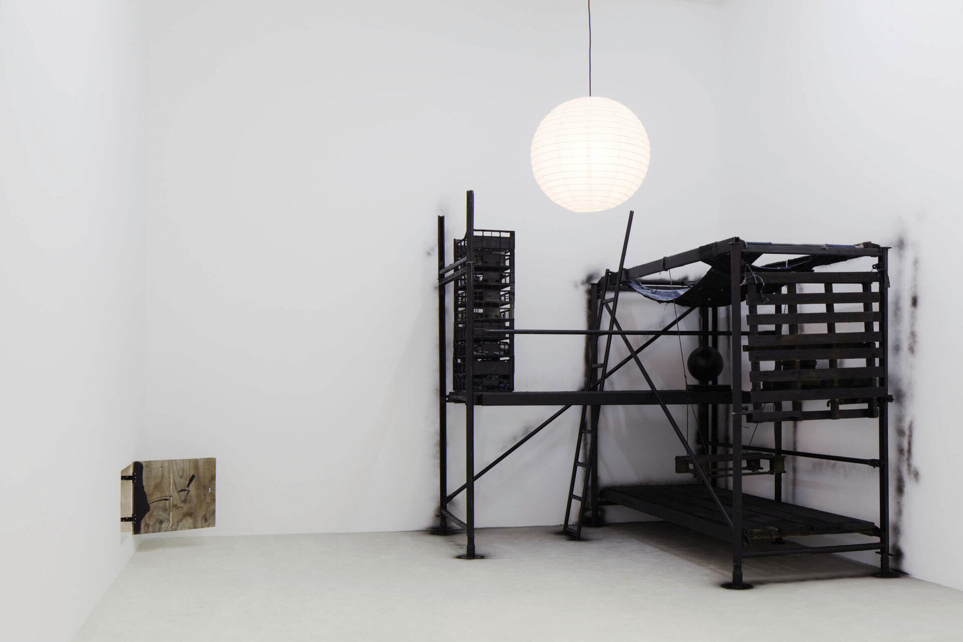 Installation von Klara Lidén mit einer schwarzen, grob gezimmerten Konstruktion aus Holzpaletten, Stahlstangen und einer Papierlampe; an der linken Wand eine aufklappbare Holzplatte mit Kritzeleien.
