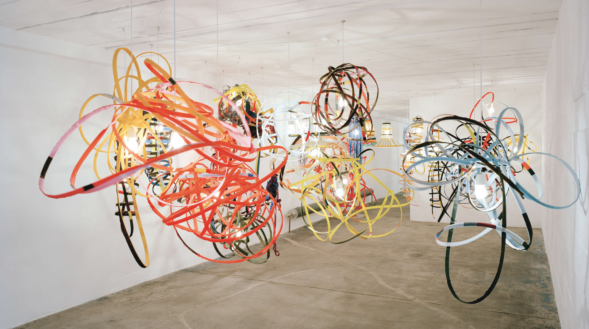 Raumfüllende Installation aus farbigen Bandstrukturen und Lampen, die von der Decke hängen, in einem weißen Raum mit Betonboden.