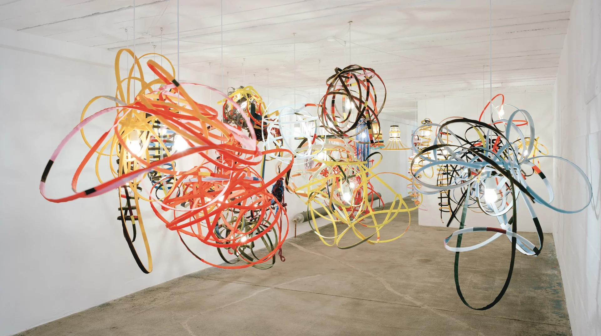 Raumfüllende Installation aus farbigen Bandstrukturen und Lampen, die von der Decke hängen, in einem weißen Raum mit Betonboden.