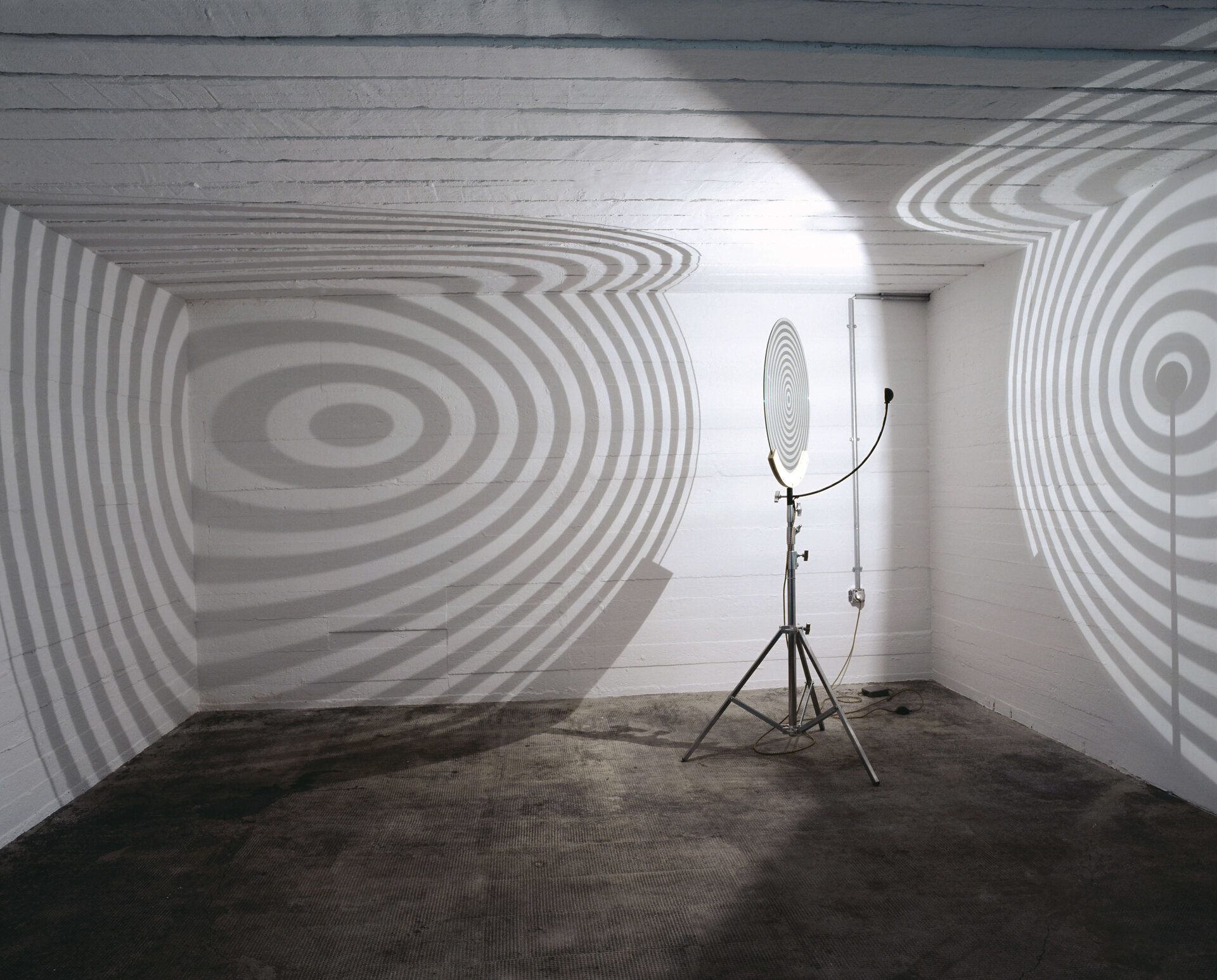 Lichtinstallation mit kreisförmiger Projektionsscheibe, die konzentrische Schattenringe an die Wände eines weißen Raumes wirft.
