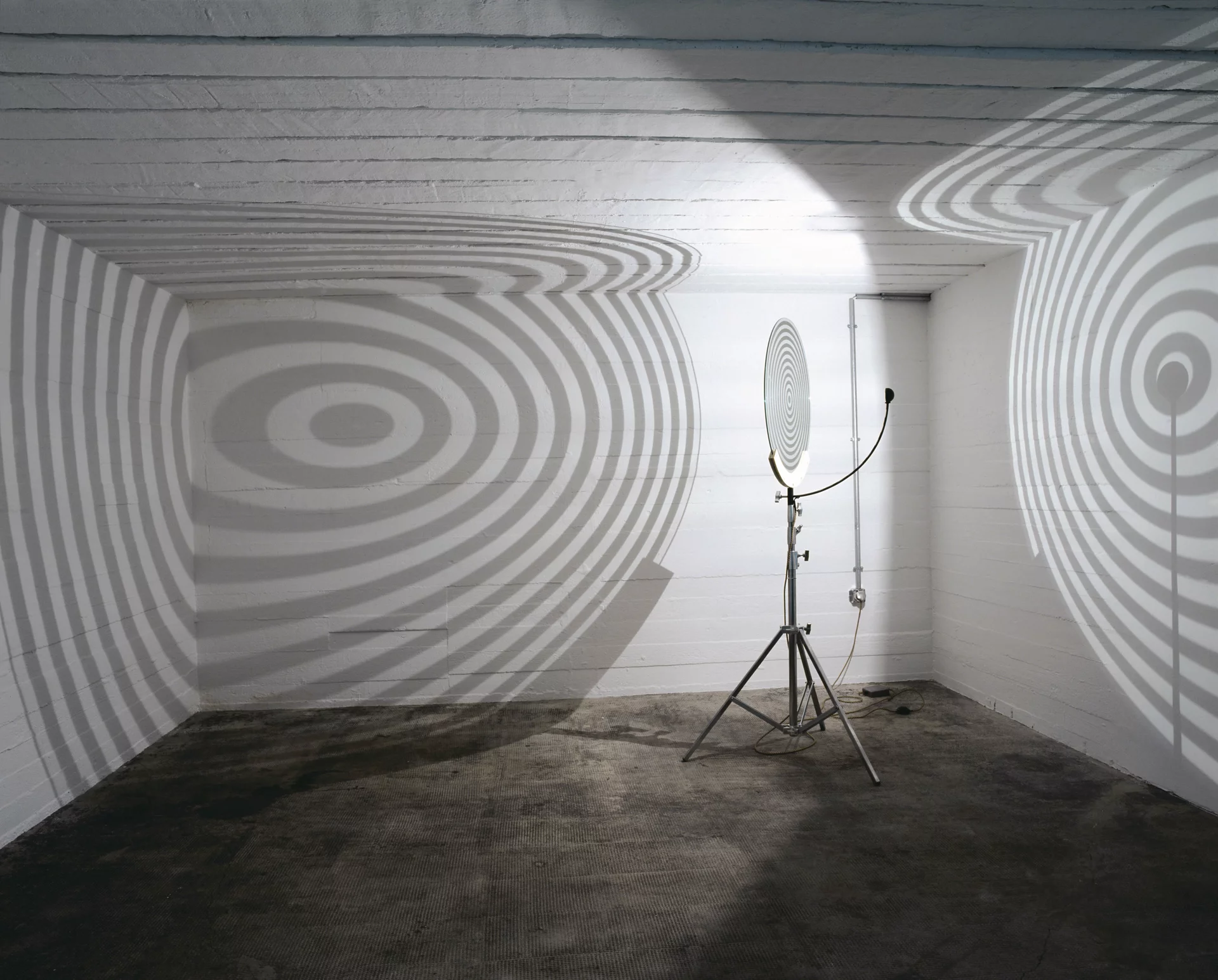 Lichtinstallation mit kreisförmiger Projektionsscheibe, die konzentrische Schattenringe an die Wände eines weißen Raumes wirft.