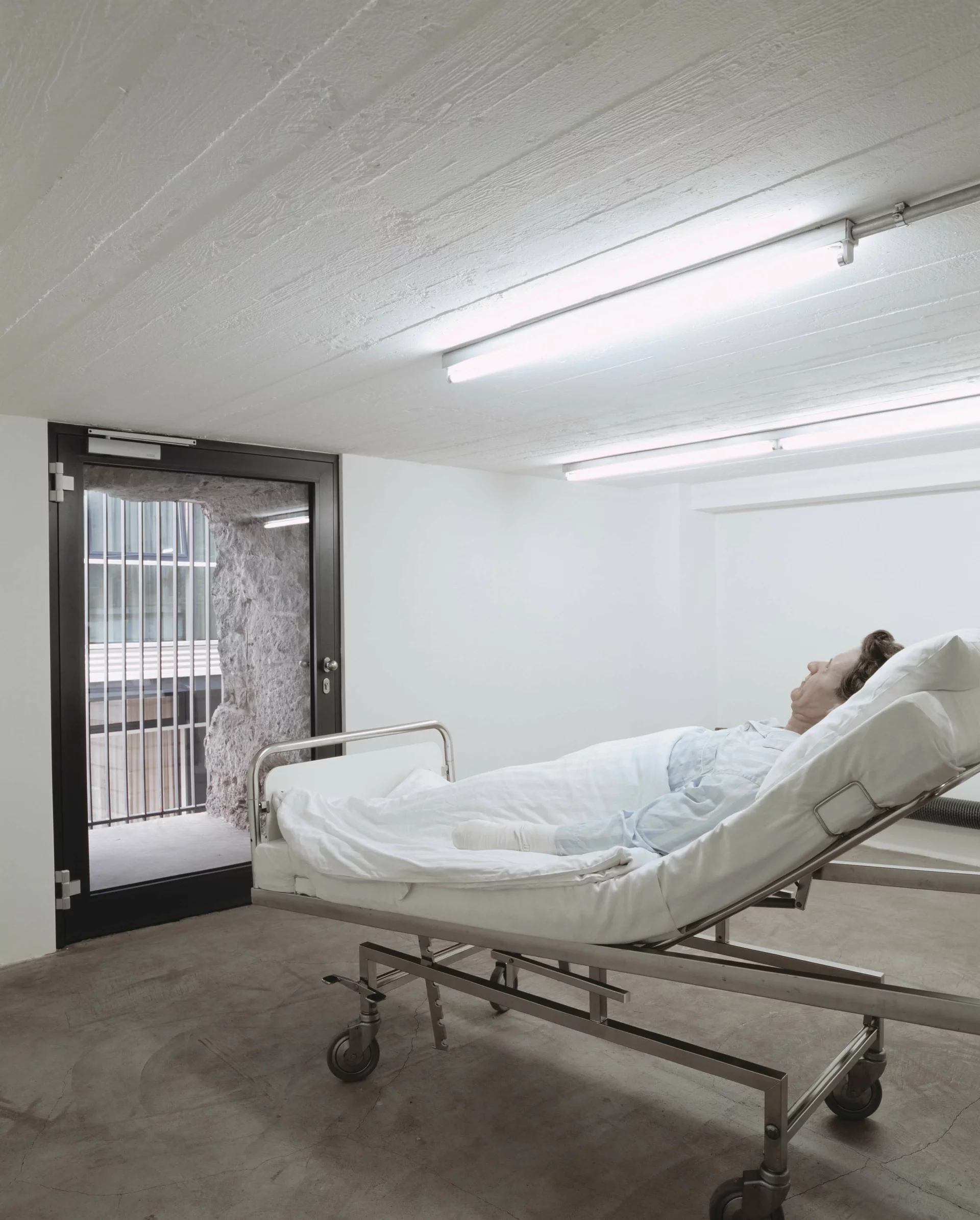Krankenbett mit lebensechter Figur in Klinikzimmer, Blick durch verglaste Tür auf Außenmauer mit Gitter.
