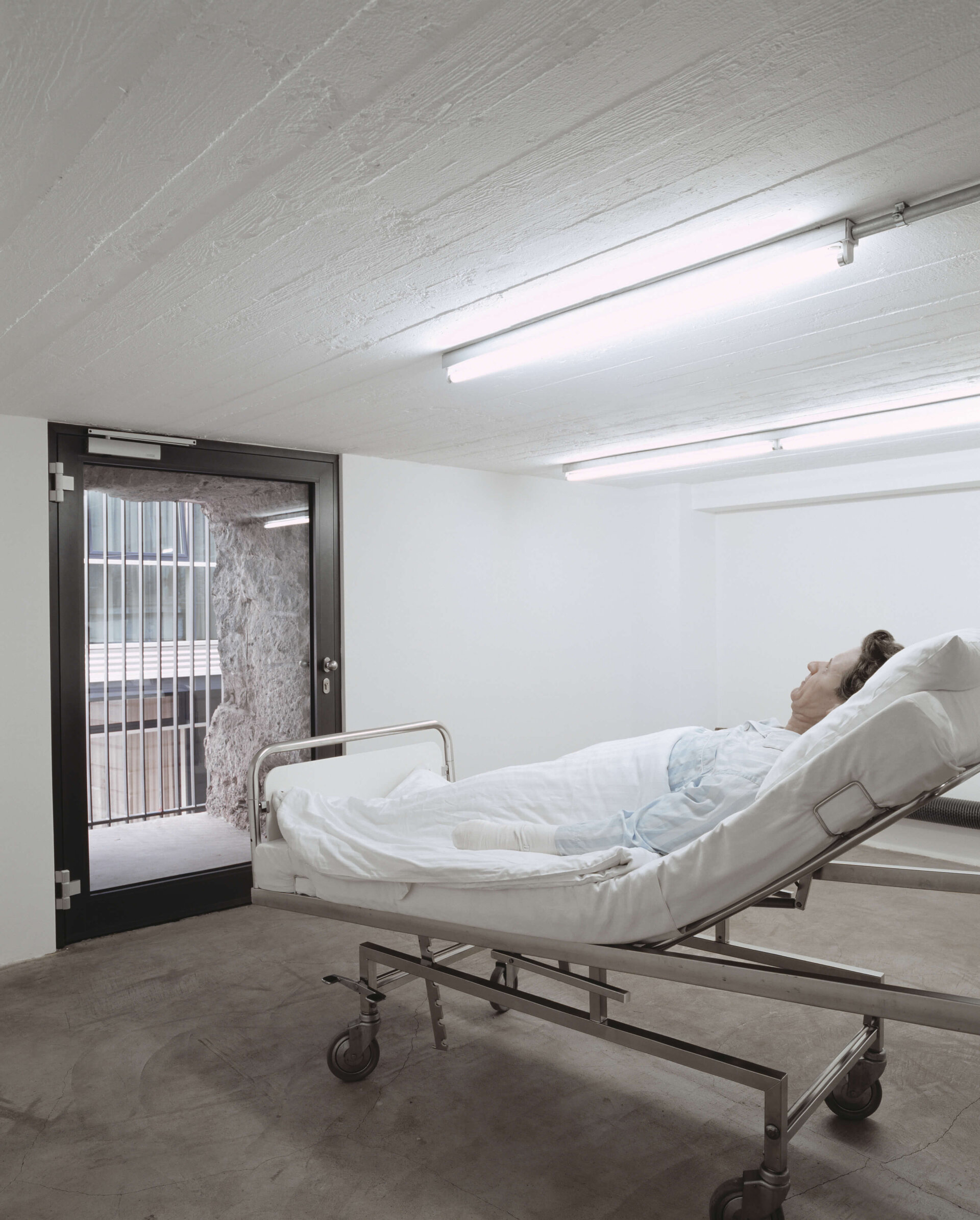 Krankenbett mit lebensechter Figur in Klinikzimmer, Blick durch verglaste Tür auf Außenmauer mit Gitter.