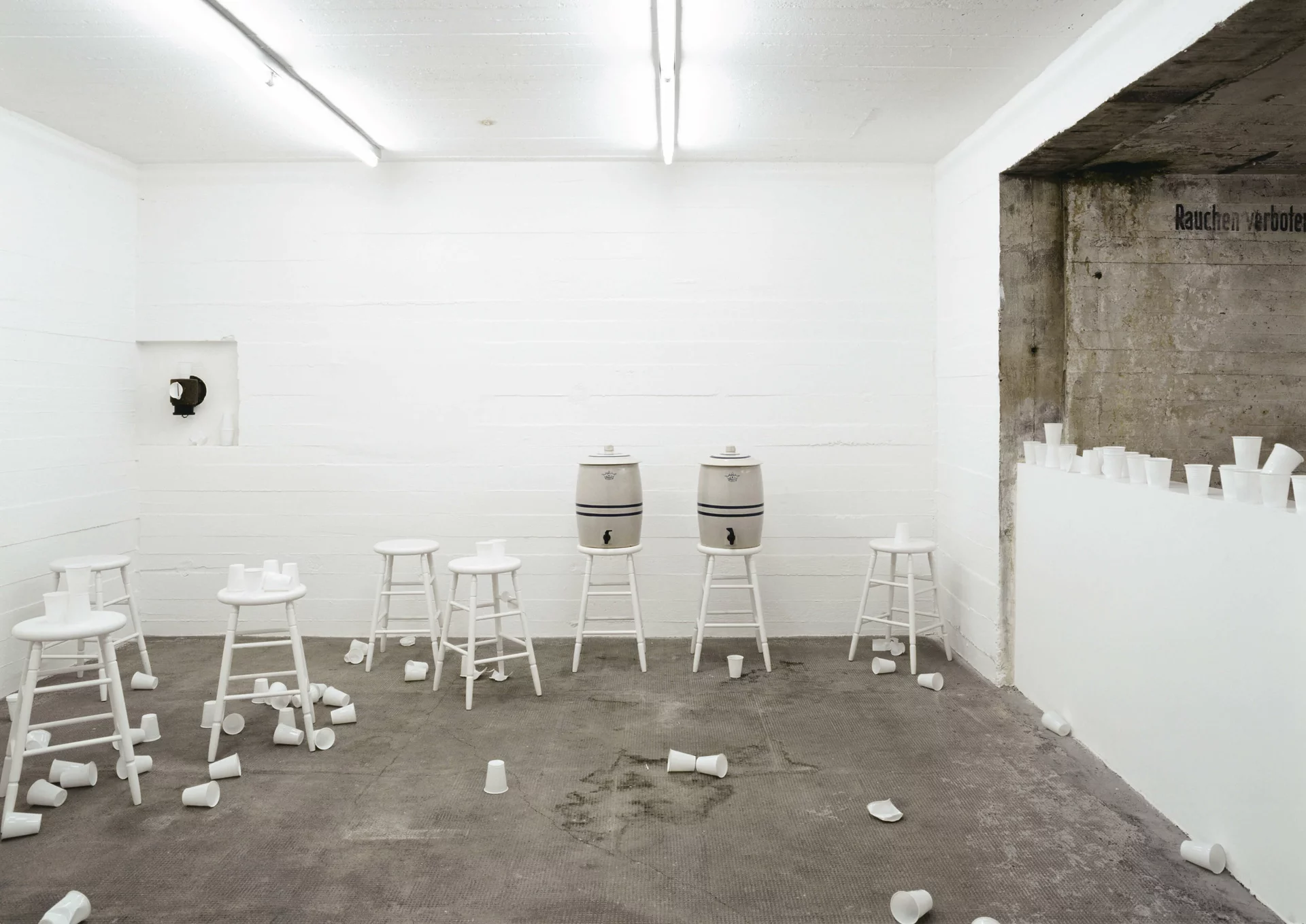 Weiße Hocker und Wasserbehälter mit verstreuten Bechern in hellem Raum; partizipative Installation von Rirkrit Tiravanija.