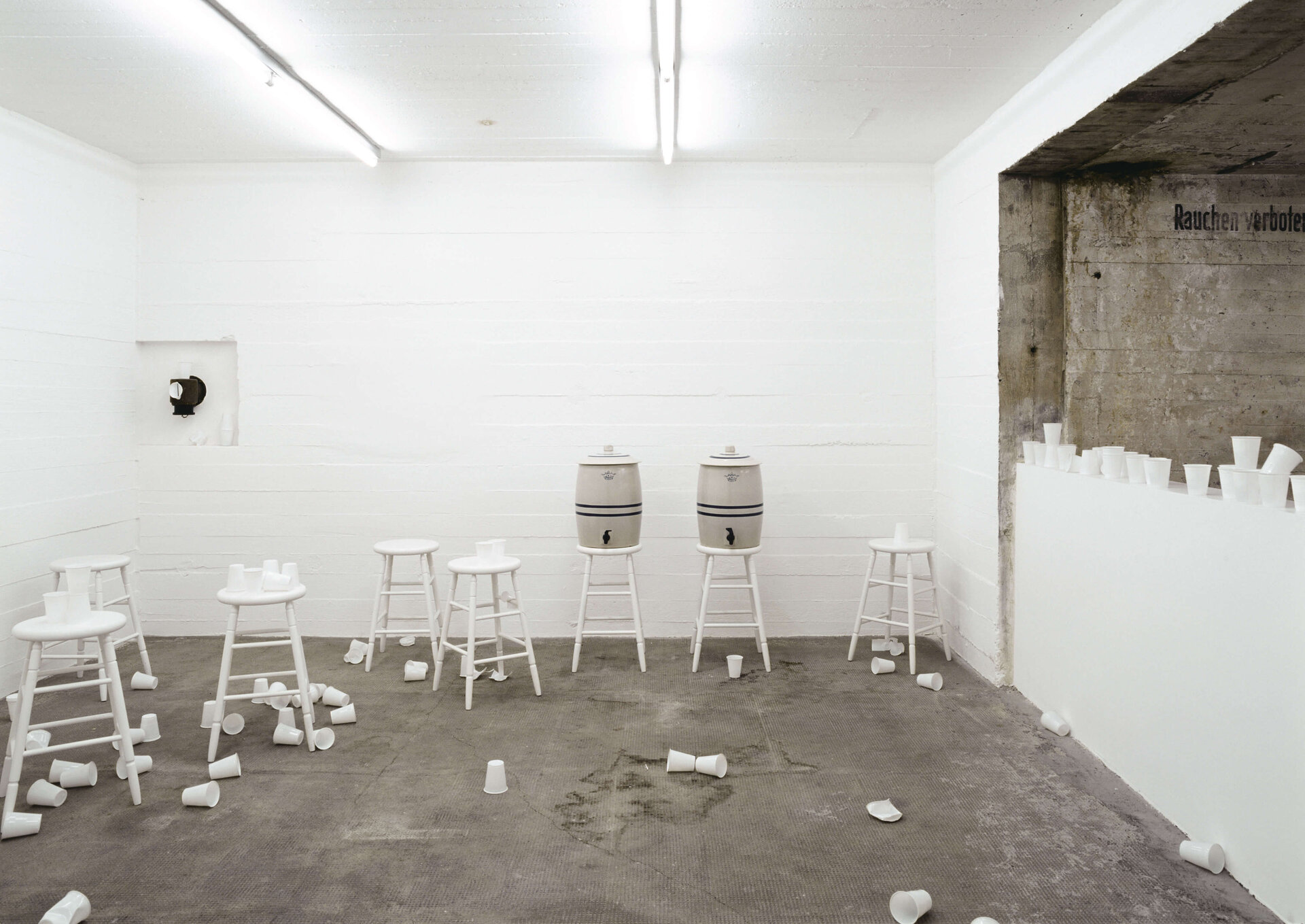 Weiße Hocker und Wasserbehälter mit verstreuten Bechern in hellem Raum; partizipative Installation von Rirkrit Tiravanija.