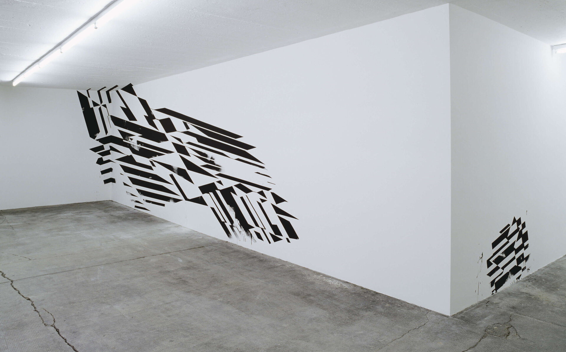 Schwarze geometrische Muster zerschneiden eine weiße Wand in einem kargen Raum des Bunkers.