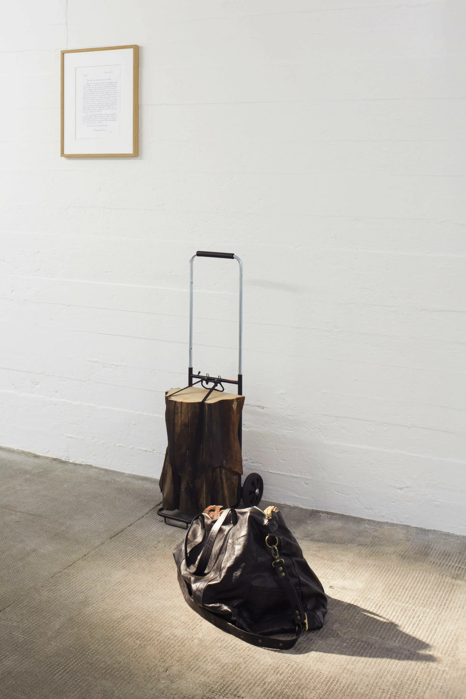 Installation von Danh Võ mit einem Holzstück auf einer Sackkarre, einer schwarzen Ledertasche am Boden und einem gerahmten handschriftlichen Brief an der weißen Wand.