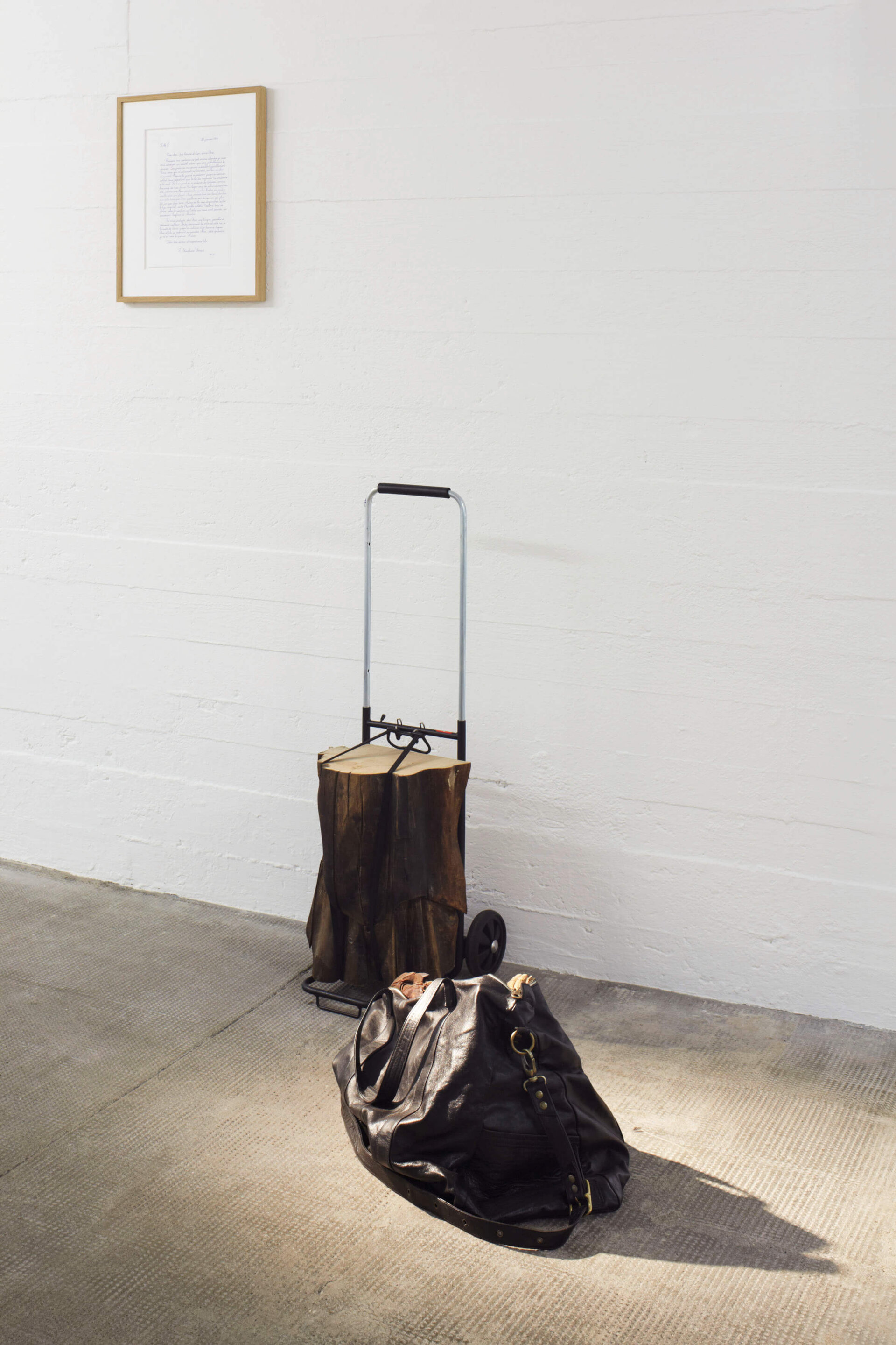 Installation von Danh Võ mit einem Holzstück auf einer Sackkarre, einer schwarzen Ledertasche am Boden und einem gerahmten handschriftlichen Brief an der weißen Wand.