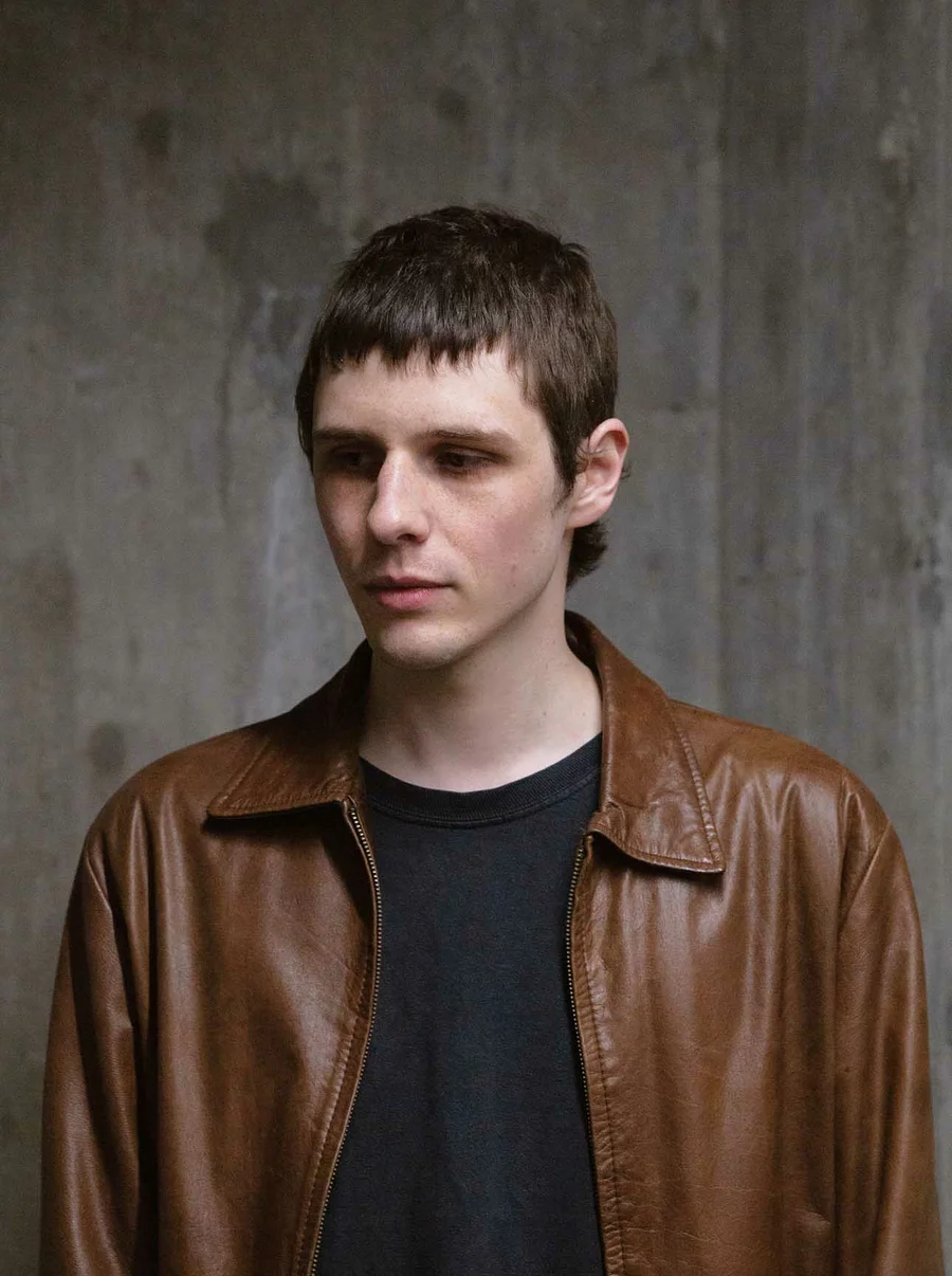 Valentin in brauner Lederjacke und schwarzem Shirt vor einer rauen Betonwand.