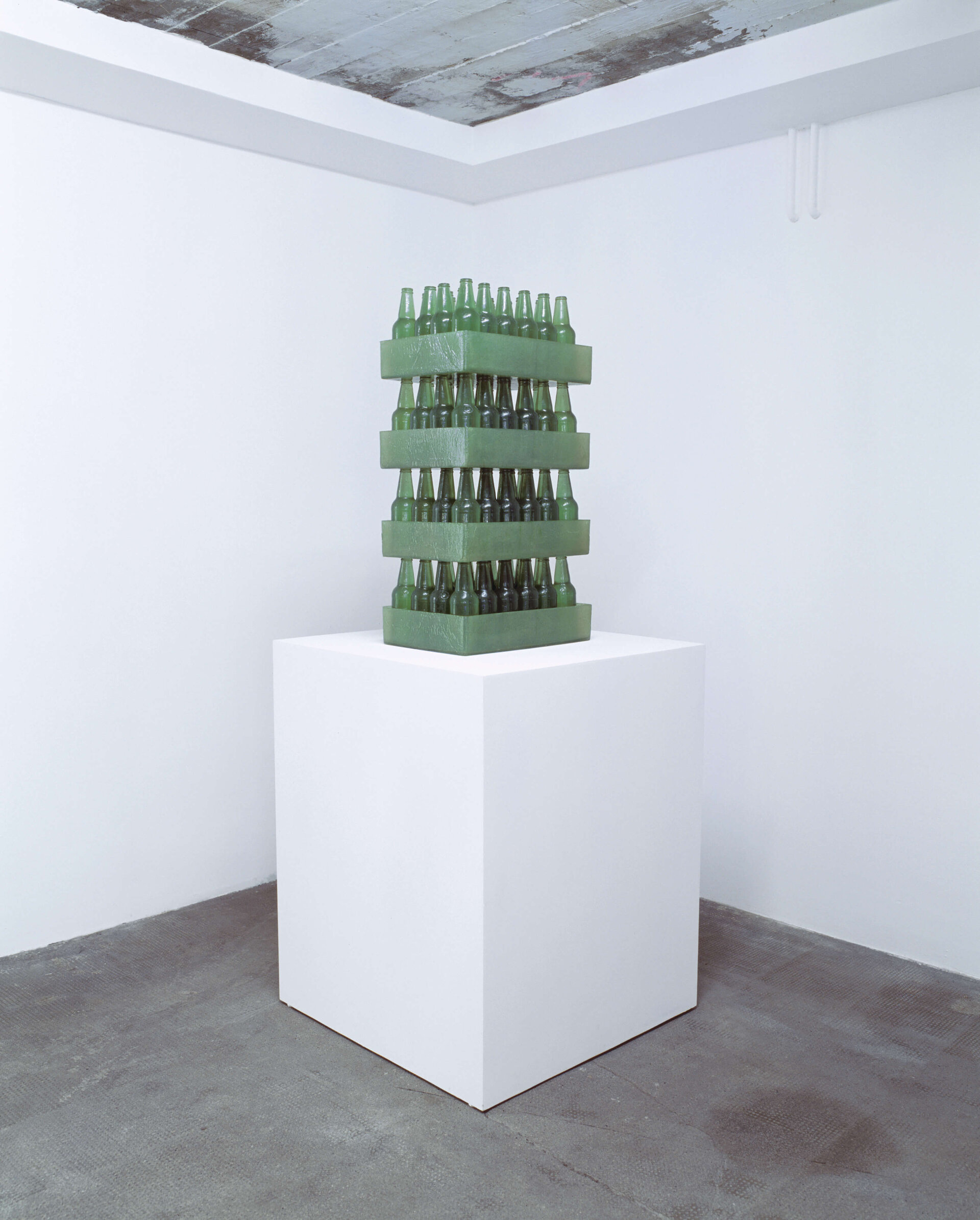 Grüne Glasflaschen in vier Getränkekisten übereinandergestapelt auf weißem Sockel in minimalistischem Ausstellungsraum.