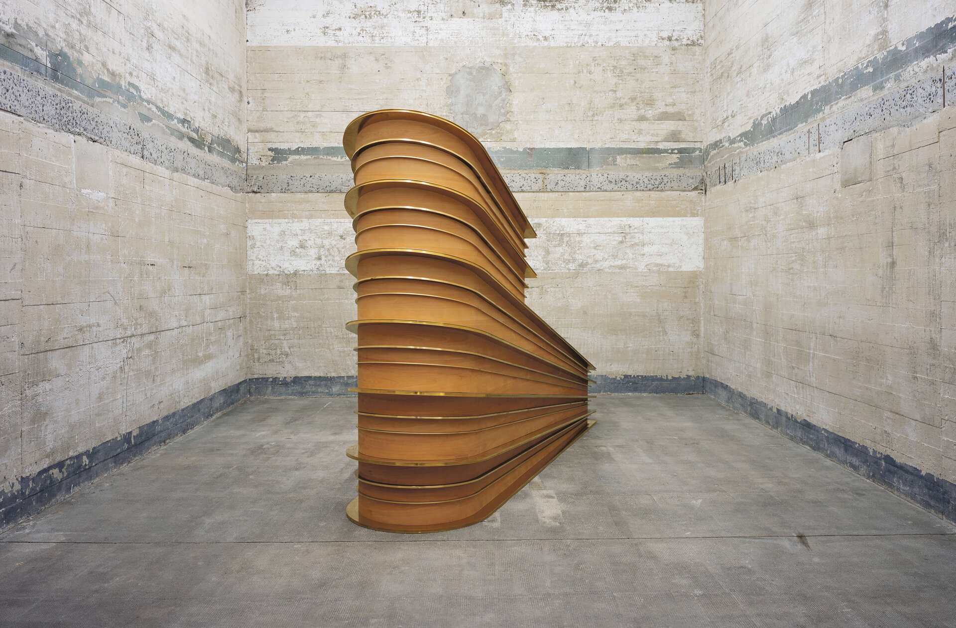 Holzskulptur mit geschichteter, gebogener Form in einem kahlen, betonierten Bunkerraum unter Neonlicht.