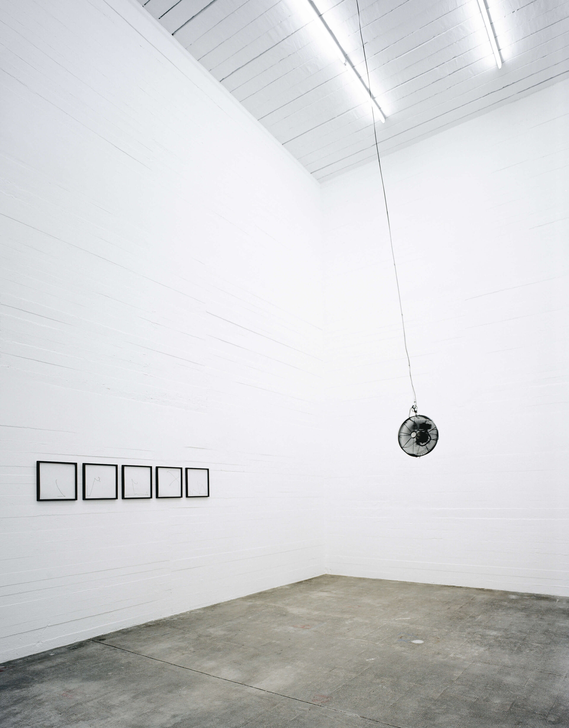 Schwarzer Ventilator hängt an Kabel von der Decke in weißem Raum mit fünf gerahmten Zeichnungen an der Wand.