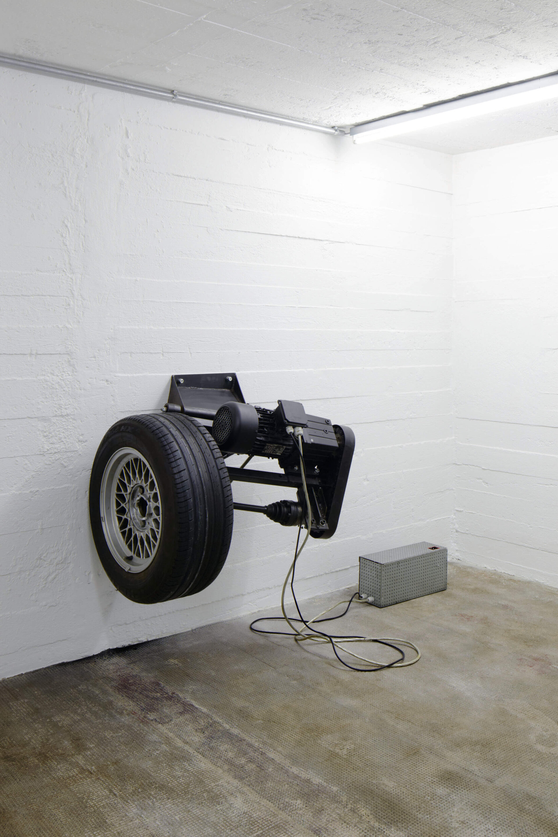 Ein drehender Autoreifen, horizontal an einer weißen Wand montiert, hinterlässt schwarze Abriebspuren auf der Wand – eine Installation von Michael Sailstorfer.