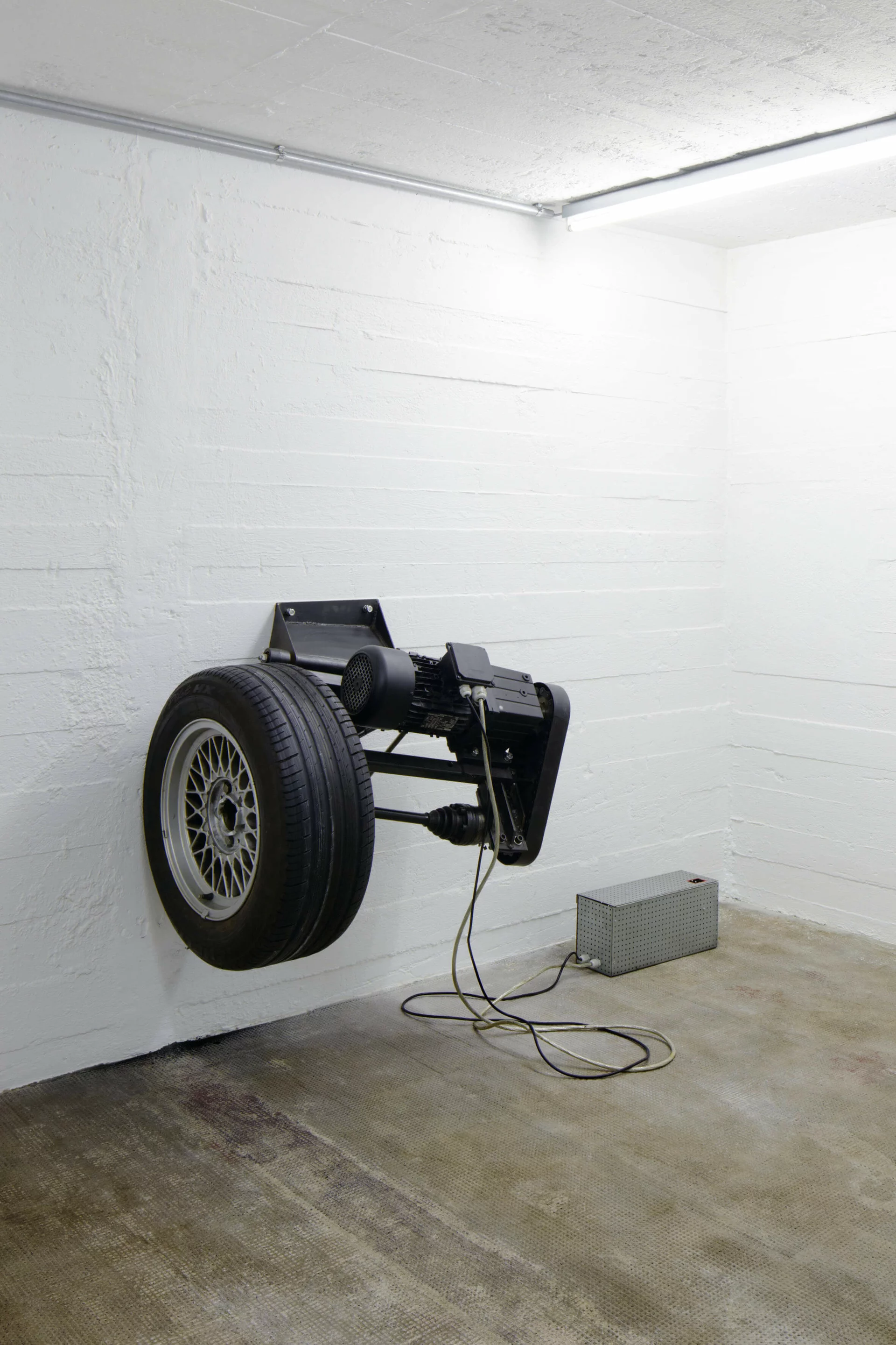 Ein drehender Autoreifen, horizontal an einer weißen Wand montiert, hinterlässt schwarze Abriebspuren auf der Wand – eine Installation von Michael Sailstorfer.