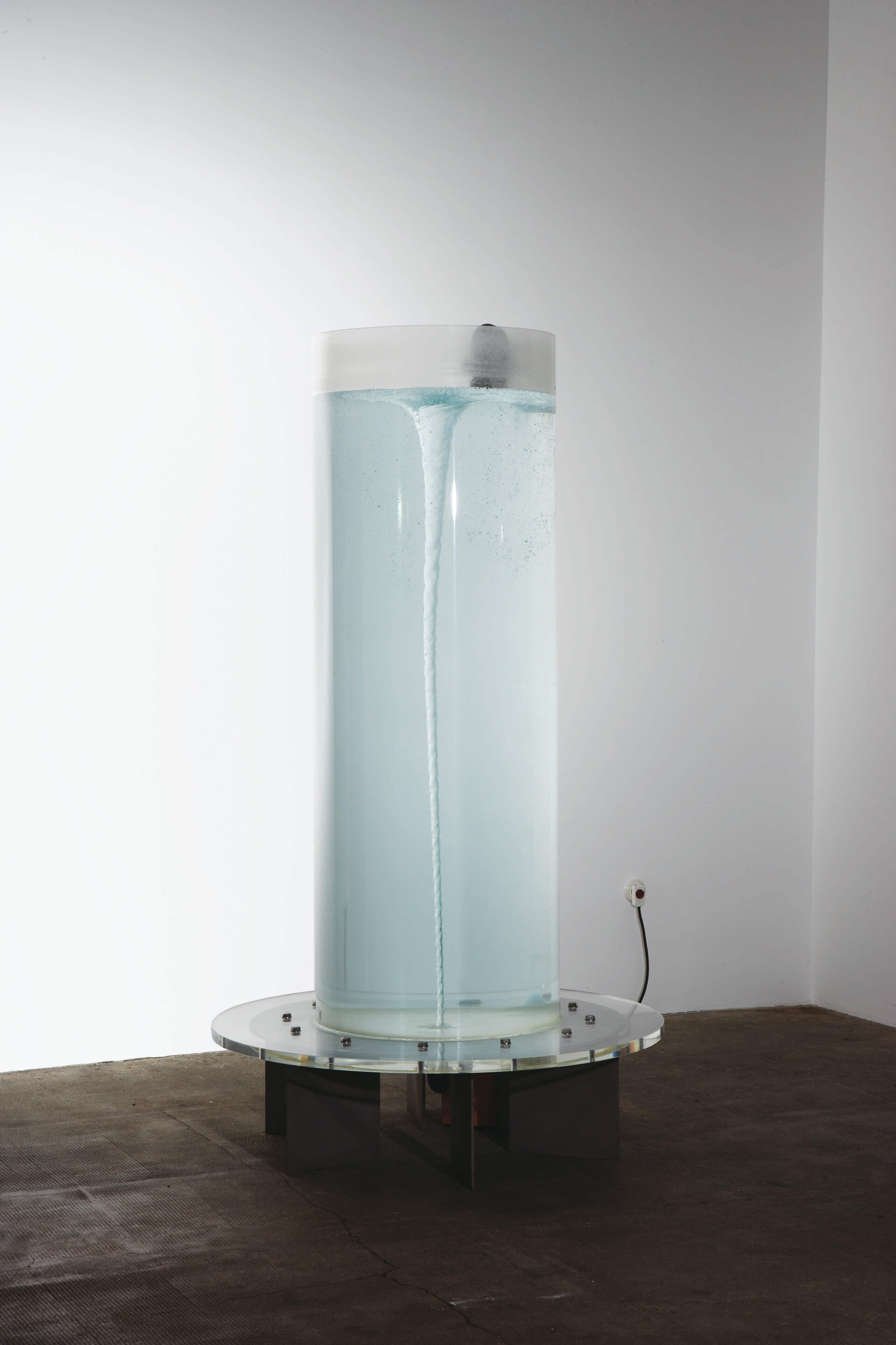 Zylindrischer Wassertank zeigt sichtbaren Wasserwirbel im Inneren, montiert auf Metallgestell in weißem Raum.