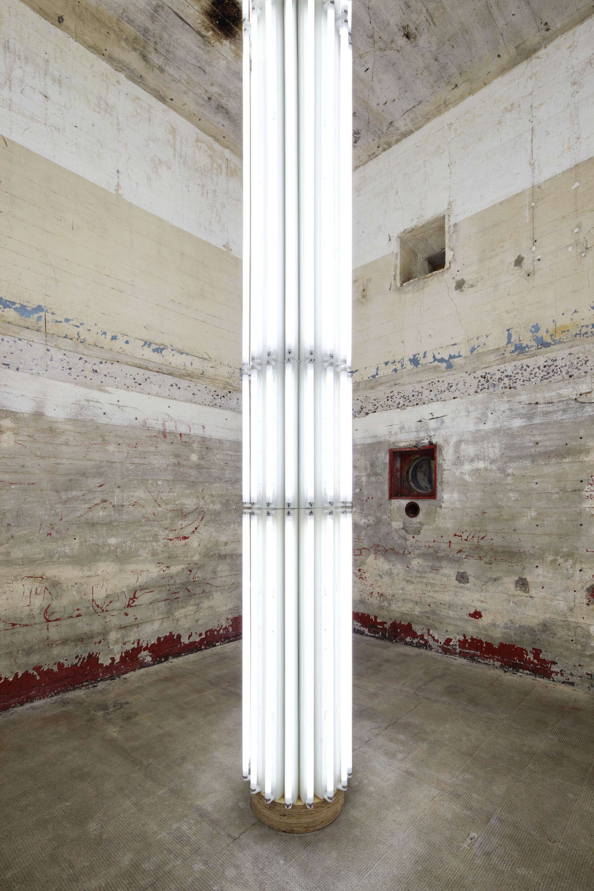 Leuchtstoffröhren-Installation von Cerith Wyn Evans in einem Betonraum mit abblätternder Wandfarbe; vertikal angeordnete, weiße Leuchtröhren formen eine skulpturale Lichtsäule.