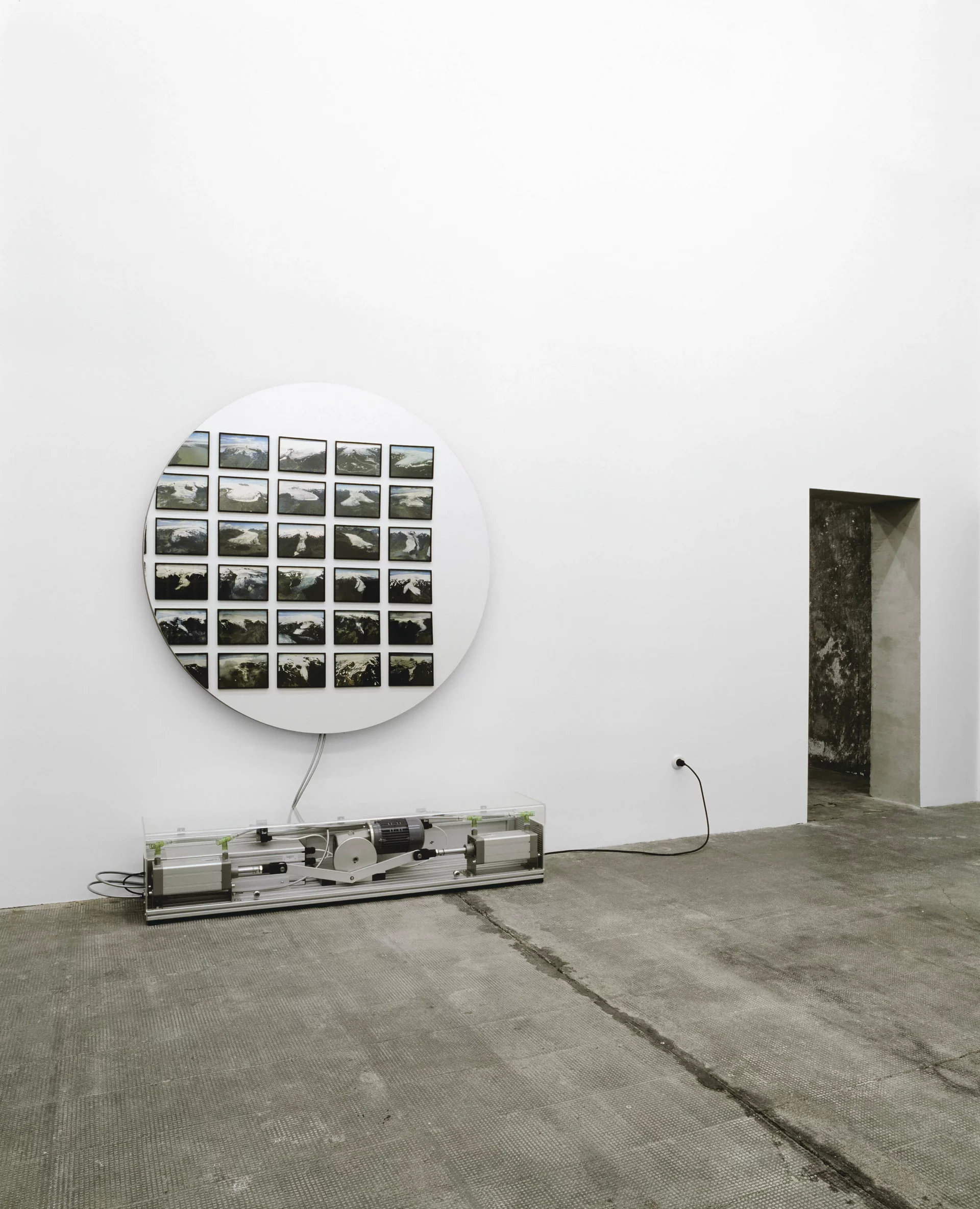Runde Fotoinstallation mit Gletscherbildern, darunter ein mechanisches Gerät in Glasvitrine an weißer Galeriewand.