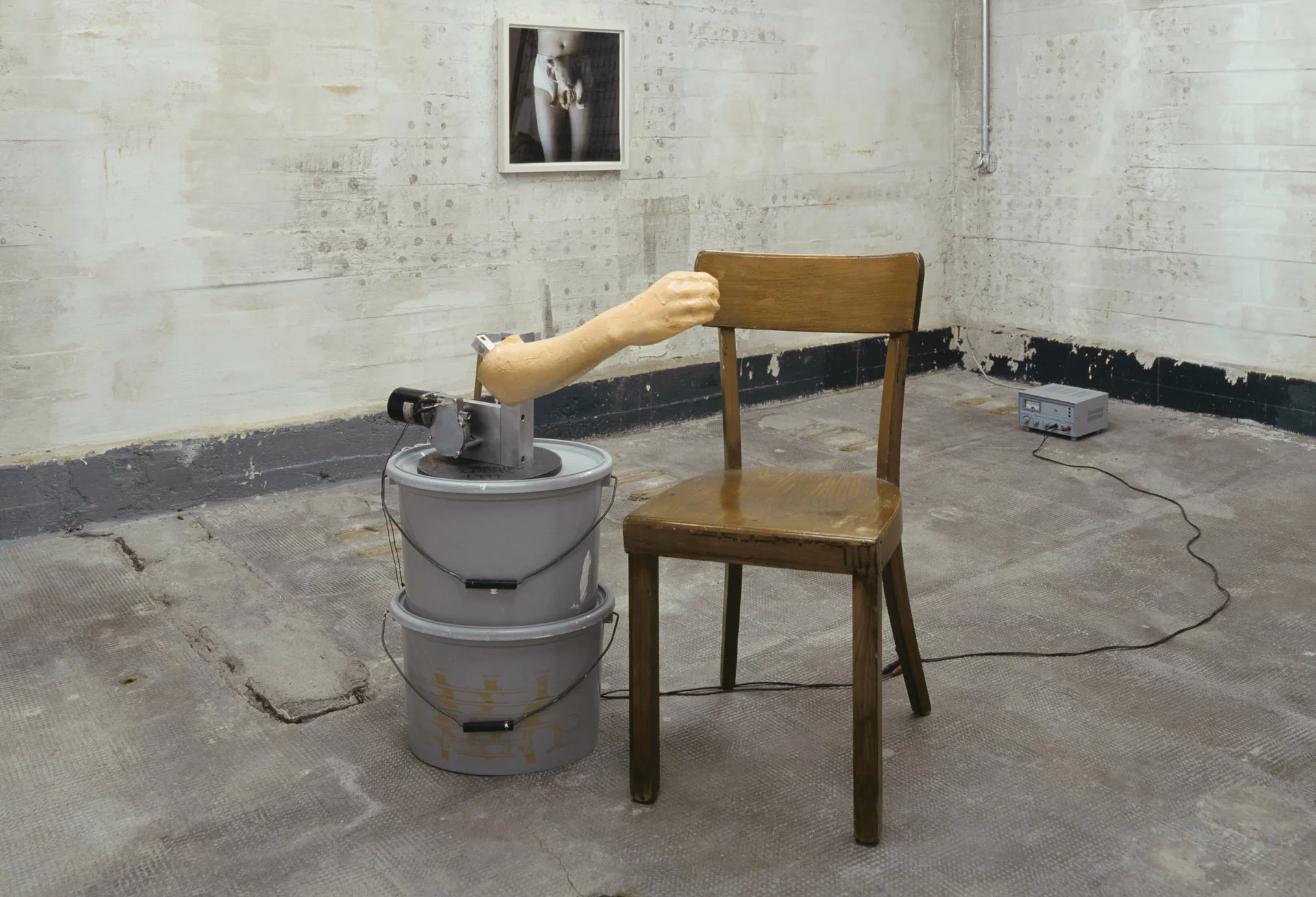 Holzstuhl neben mechanischem Arm auf Eimern in weißem Raum mit Foto einer Unterhose an der Wand.