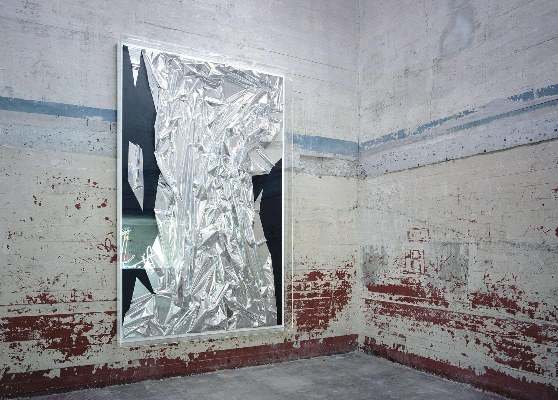 Großformatiger Spiegel mit zerknitterter Folie, reflektiert Neonlicht in einem roh belassenen Bunkerraum.