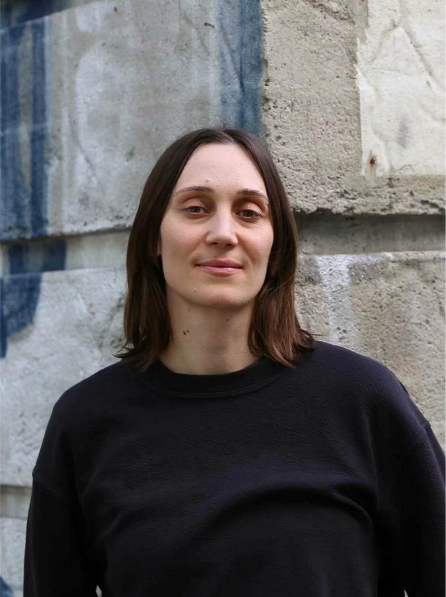 Porträt von Samantha mit schwarzem Oberteil vor einer Betonwand mit Graffiti-Elementen.