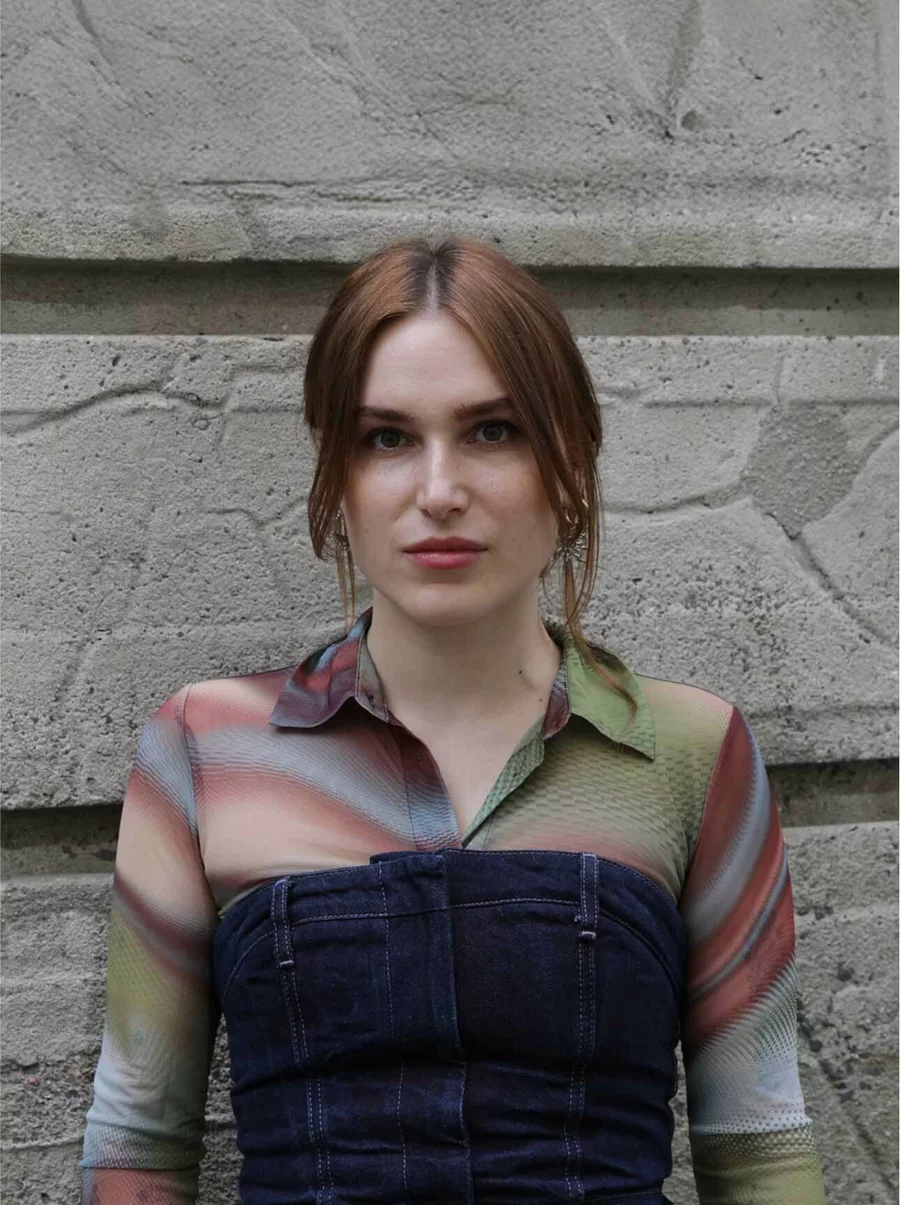 Porträt von Monya mit buntem Oberteil und offenem Blick vor strukturierter Betonwand.