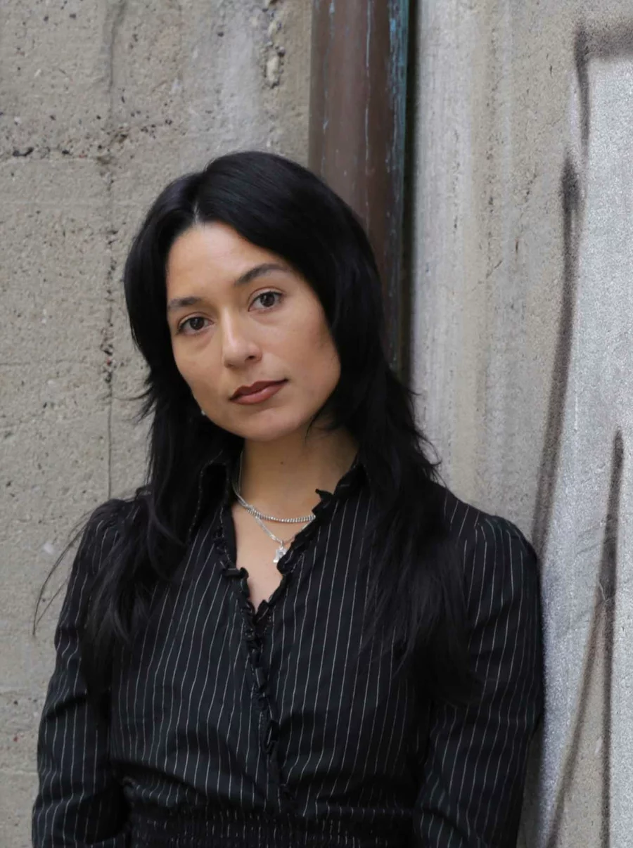 Eli vom Team der Boros Collection vor einer Betonwand mit Graffiti, in schwarzer Bluse, ruhig in die Kamera blickend.