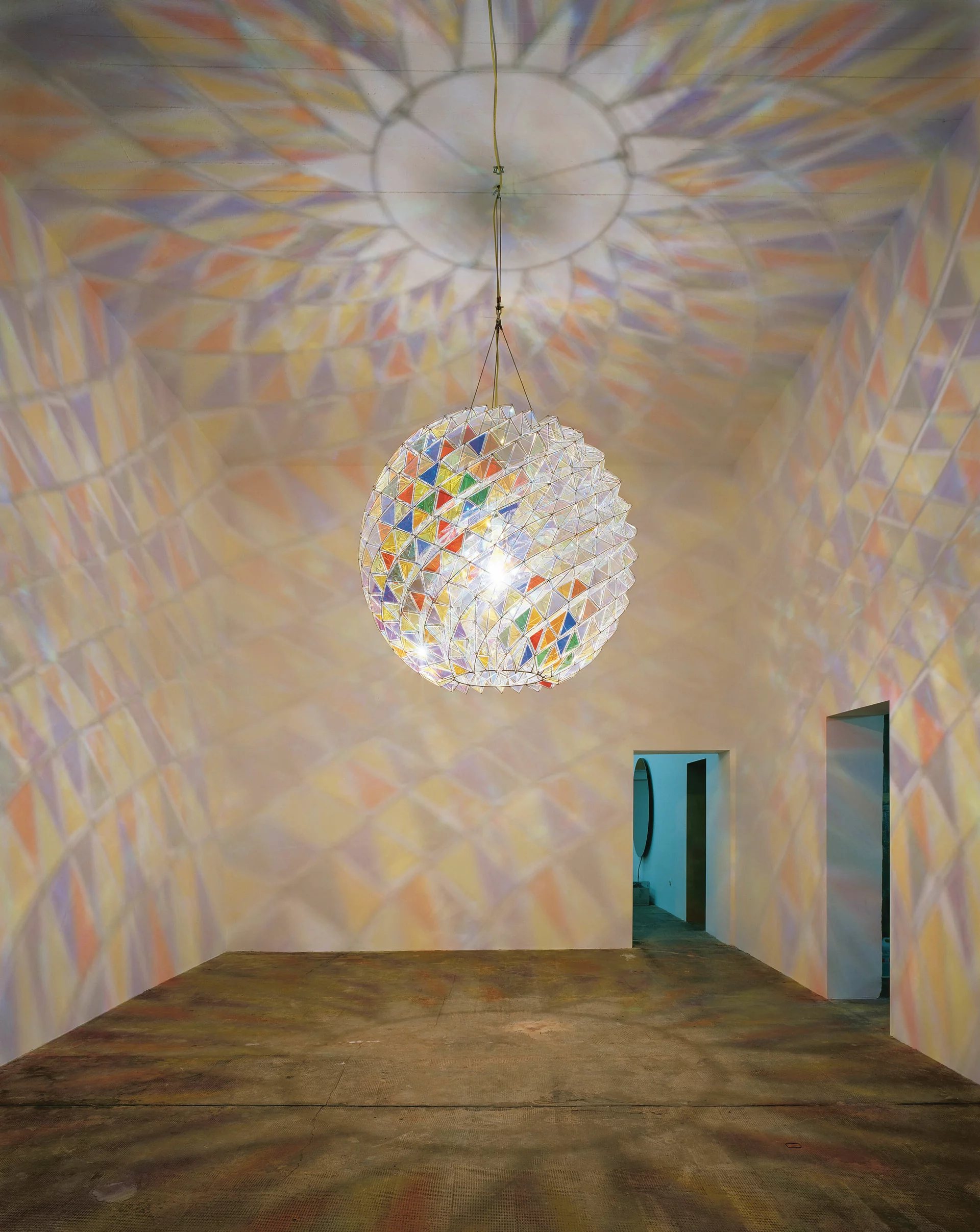 Kristallkugelartige Lichtinstallation, die bunte geometrische Muster an Wände, Decke und Boden eines weißen Raumes projiziert.