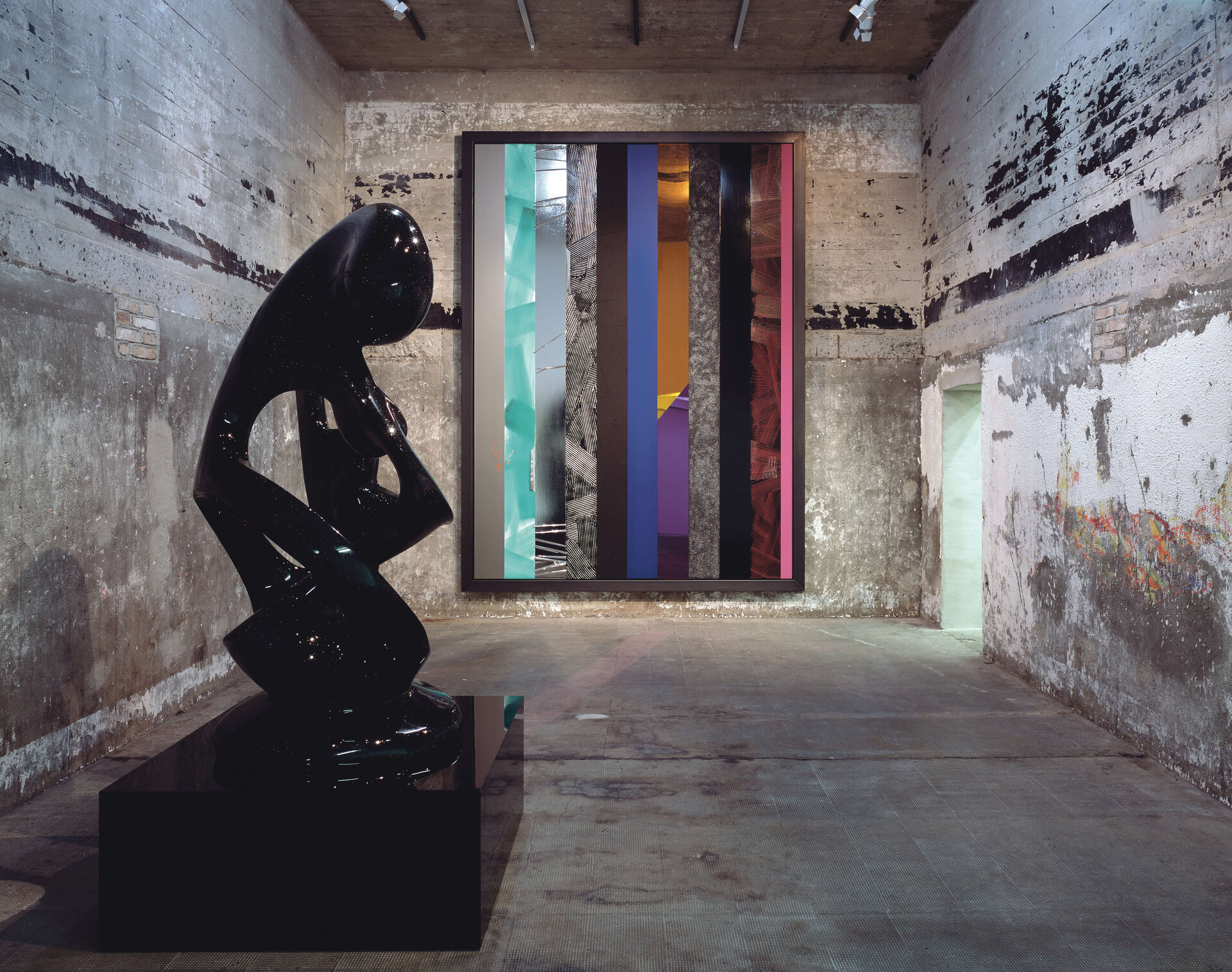 Schwarze, glänzende Skulptur vor farbigem Streifenbild in einem rauen Betonraum.