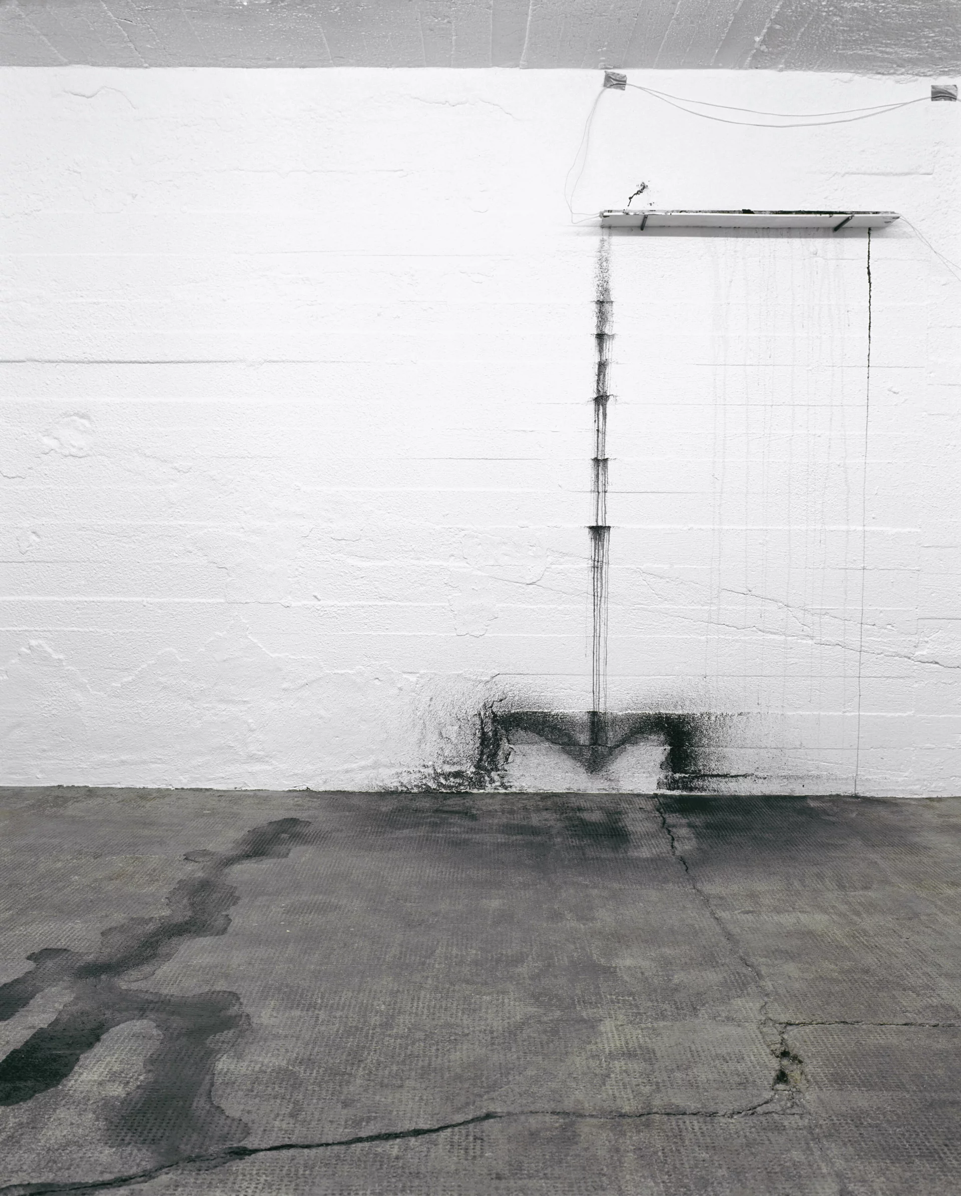 Installation an einer weißen Wand mit verlaufener schwarzer Tusche, die Spuren auf Boden und Mauer hinterlässt.
