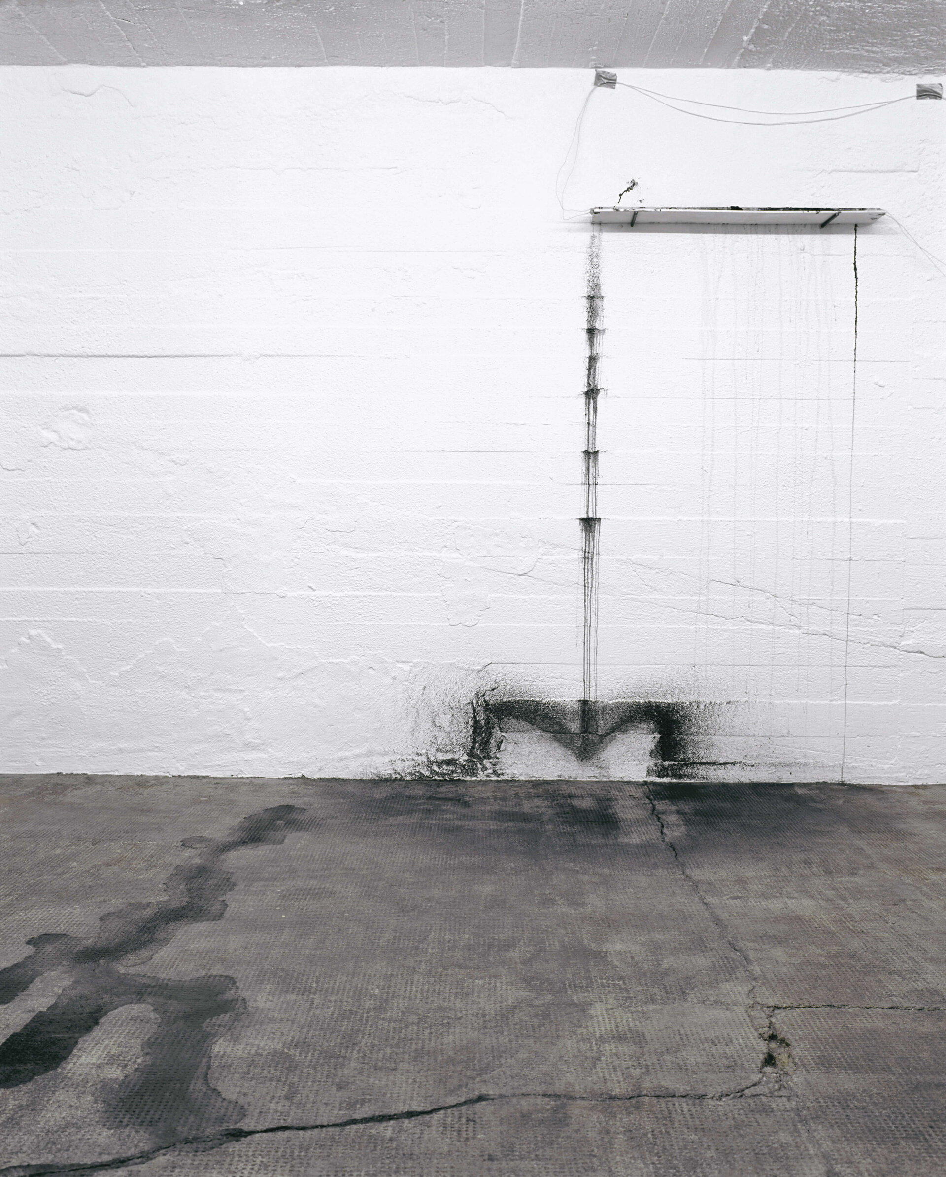 Installation an einer weißen Wand mit verlaufener schwarzer Tusche, die Spuren auf Boden und Mauer hinterlässt.