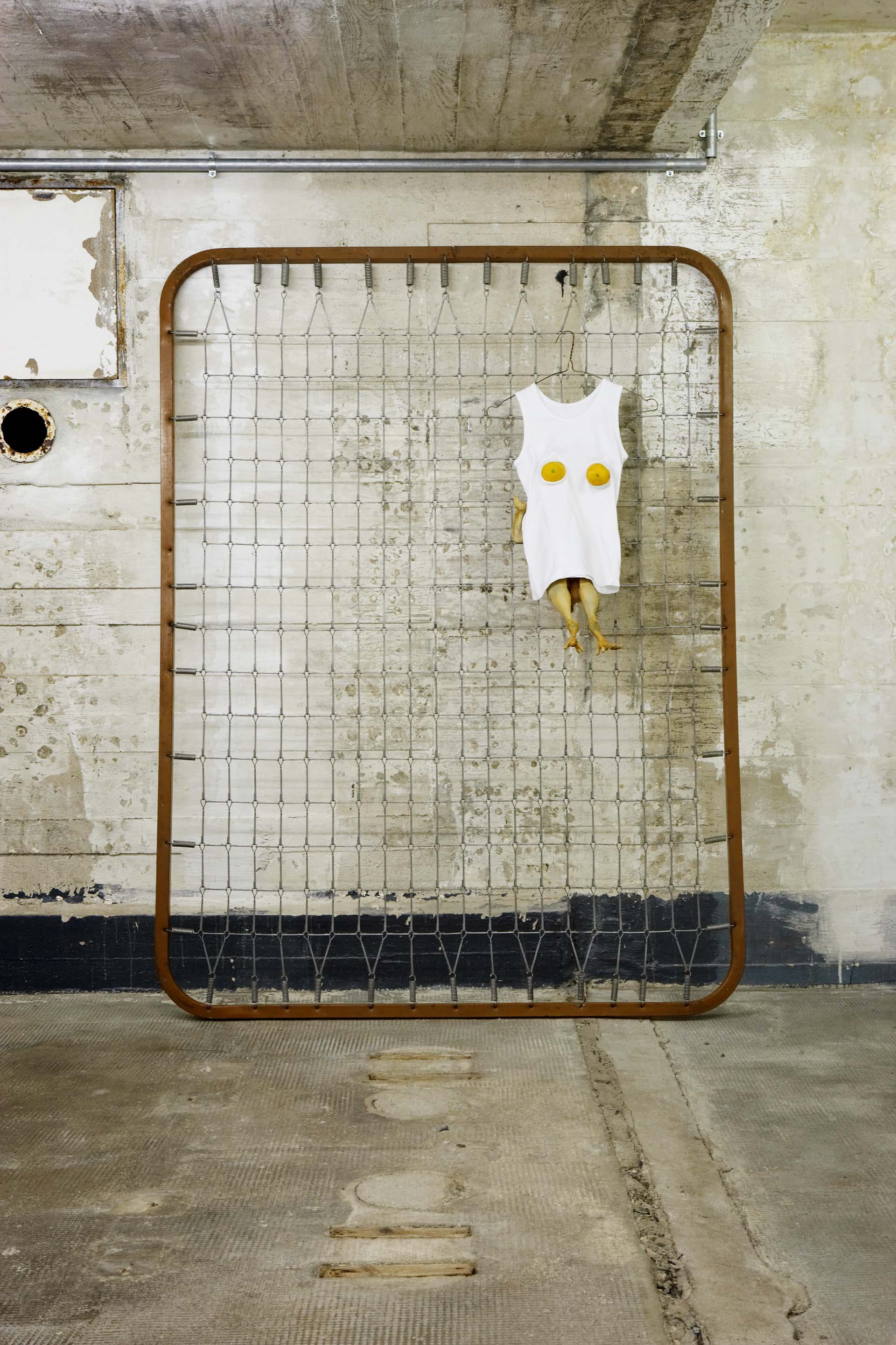 Matratzengestell an Wand gelehnt, darauf ein weißes Top mit Zitronen und darunterhängenden Gummihähnchen.